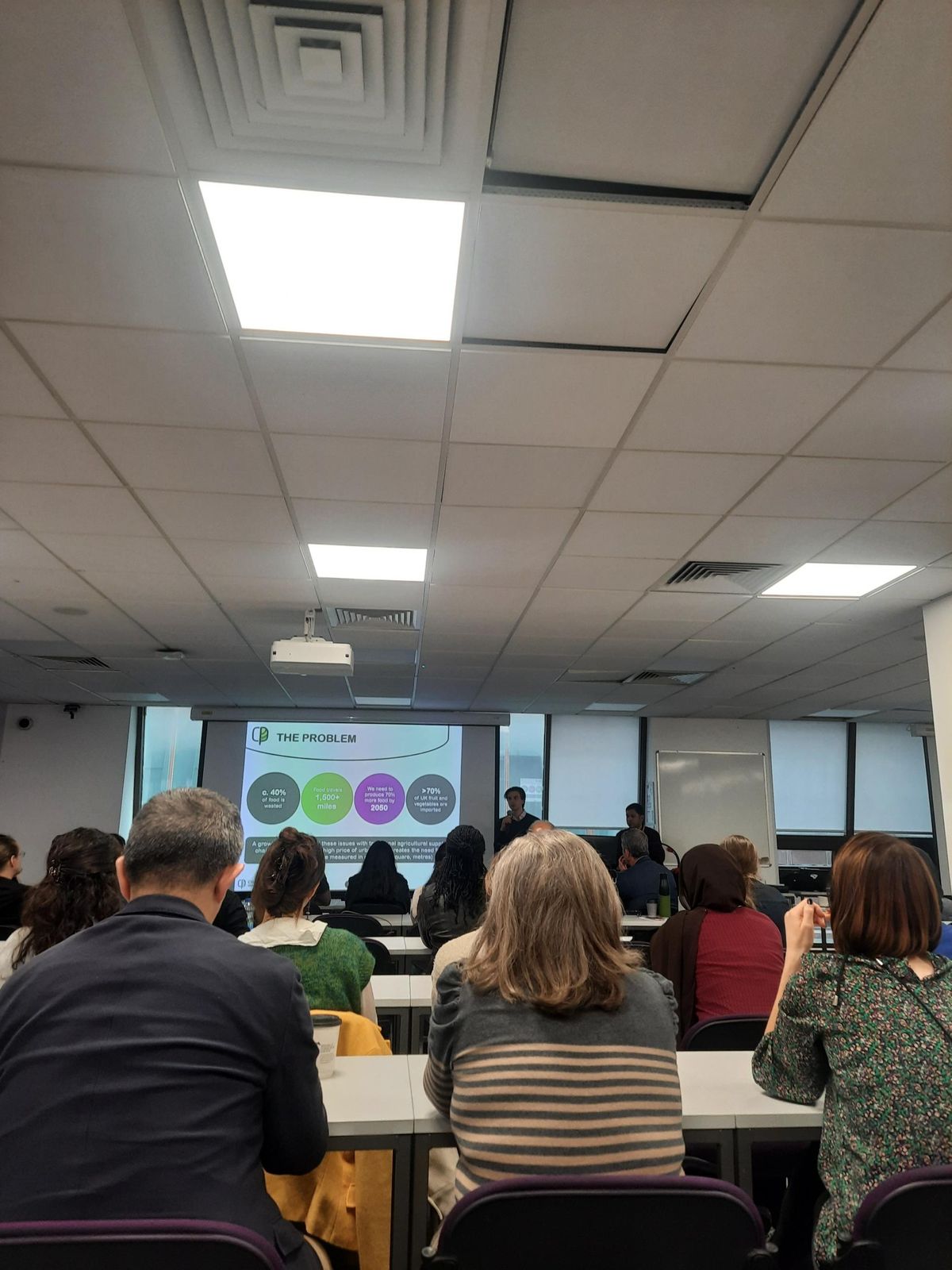 Audience in a seminar room facing a speaker and a slide titled The Problem about food waste and imported produce