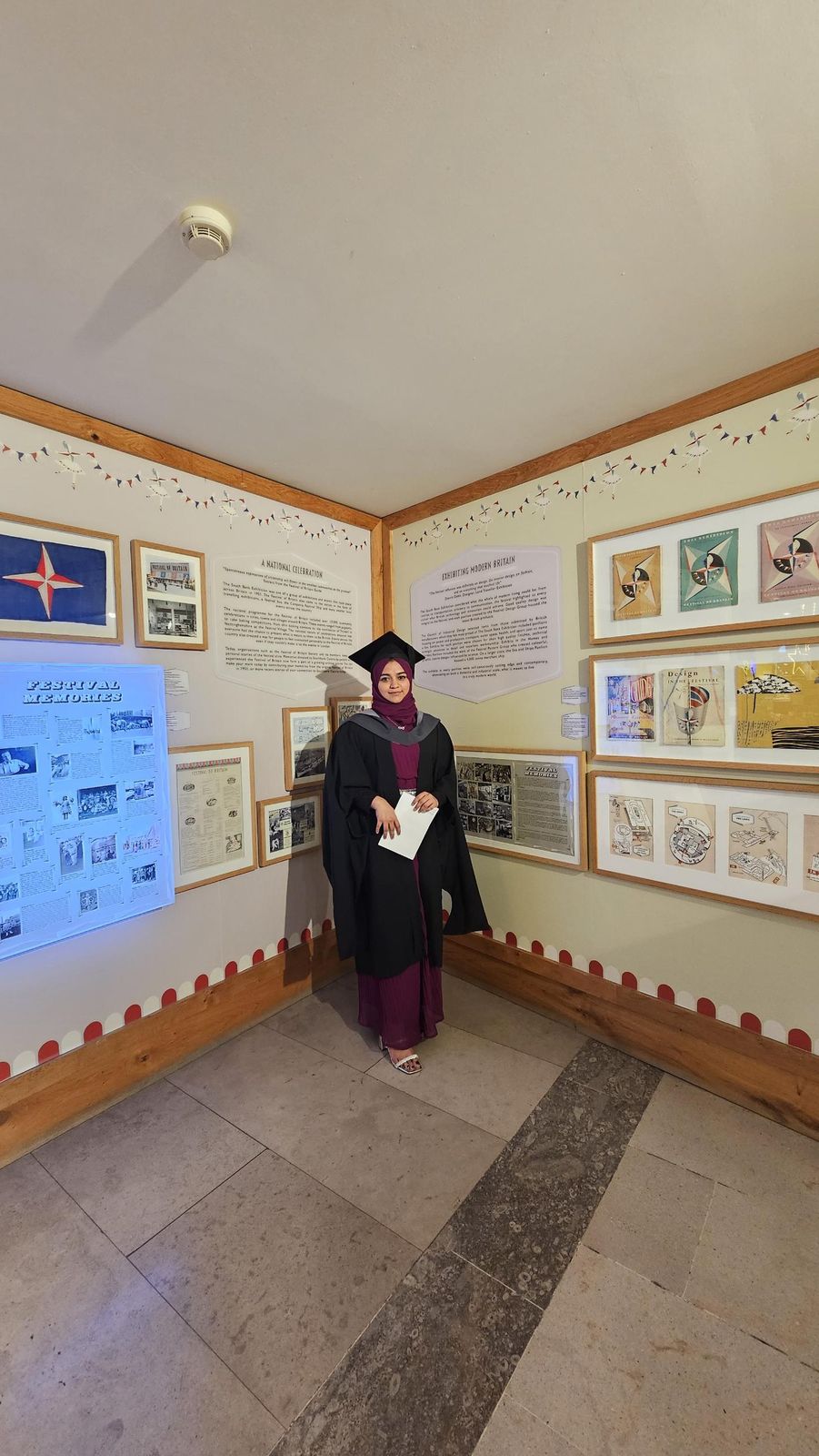 Subaiba in graduation gown and cap standing in an exhibition corner about the Festival of Britain