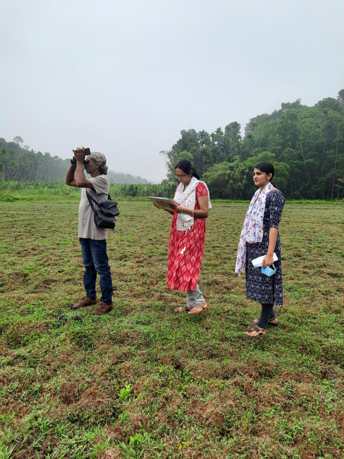 A man with binoculars and two women with clipboards standing in an open field edged by forest