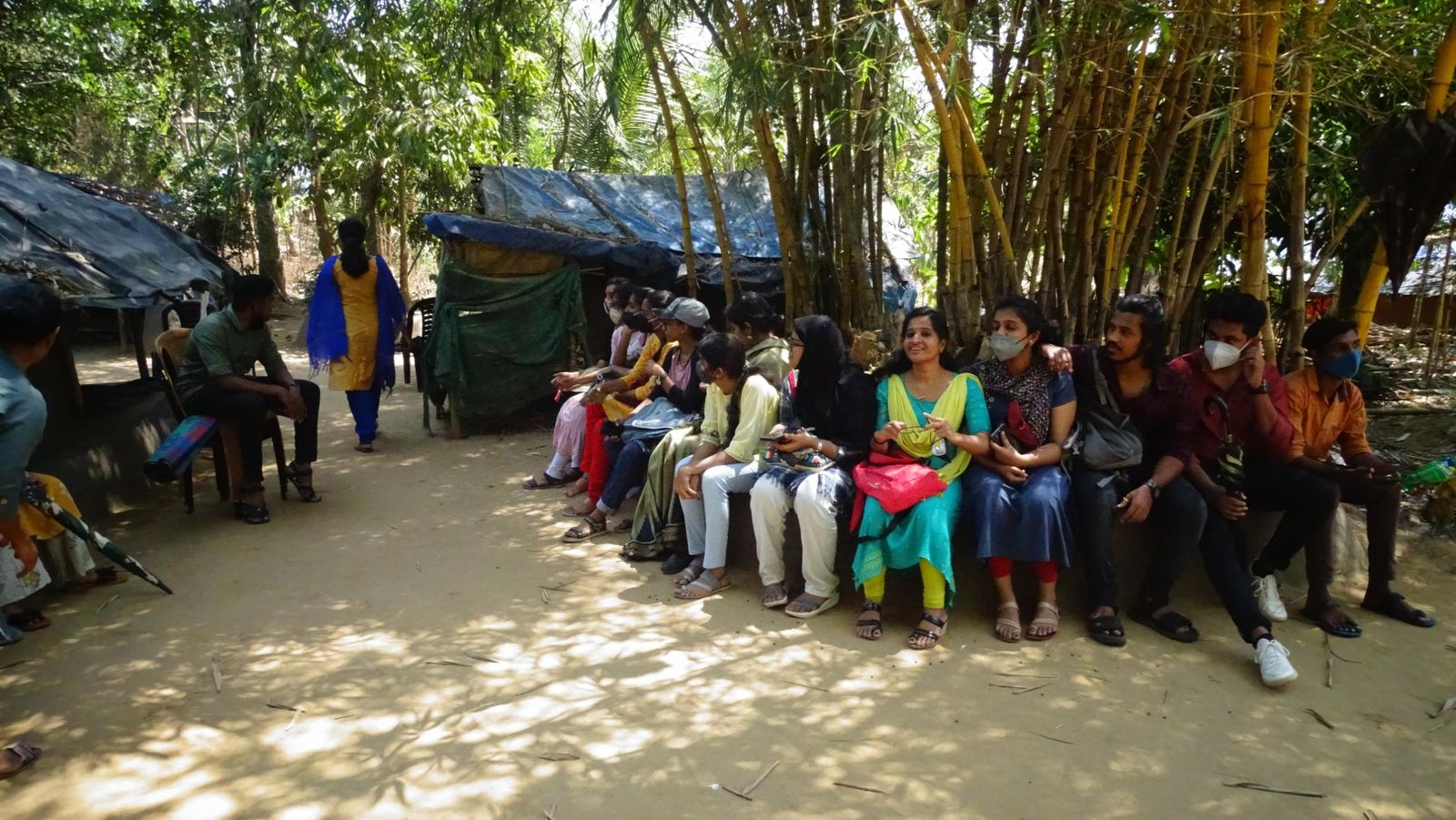 Students seated in a row under bamboo next to tarpaulin-roofed huts, in conversation with community members