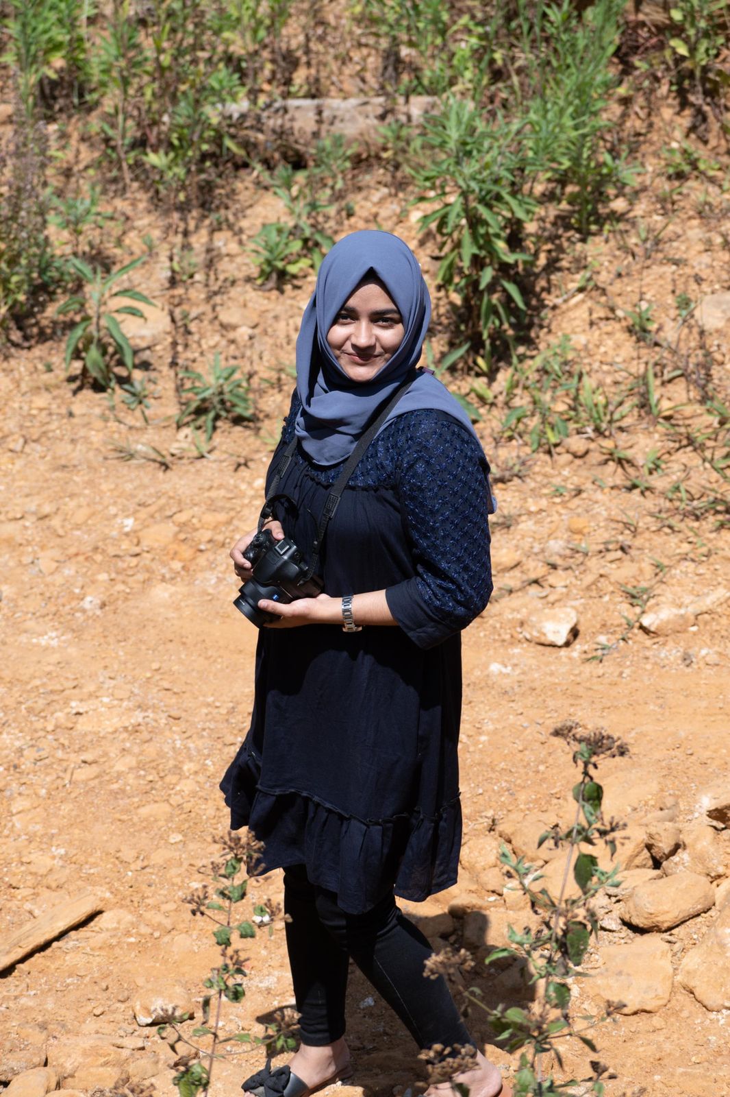 Subaiba standing on a dry hillside with a camera, scattered plants behind her