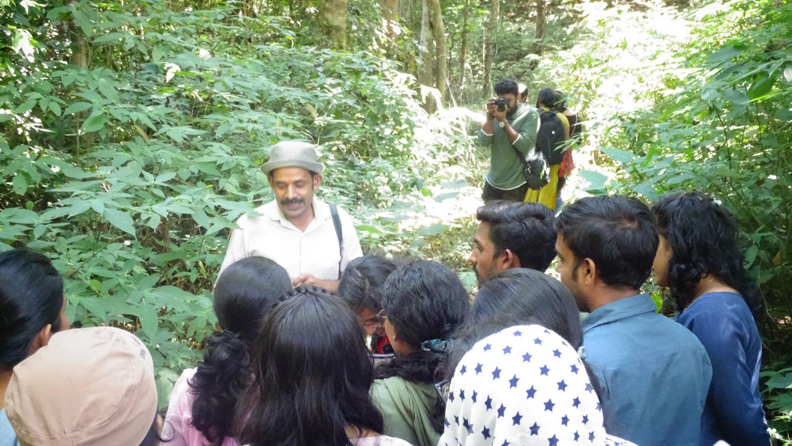 A field guide in a sun hat explaining a plant to a group of students in dense forest