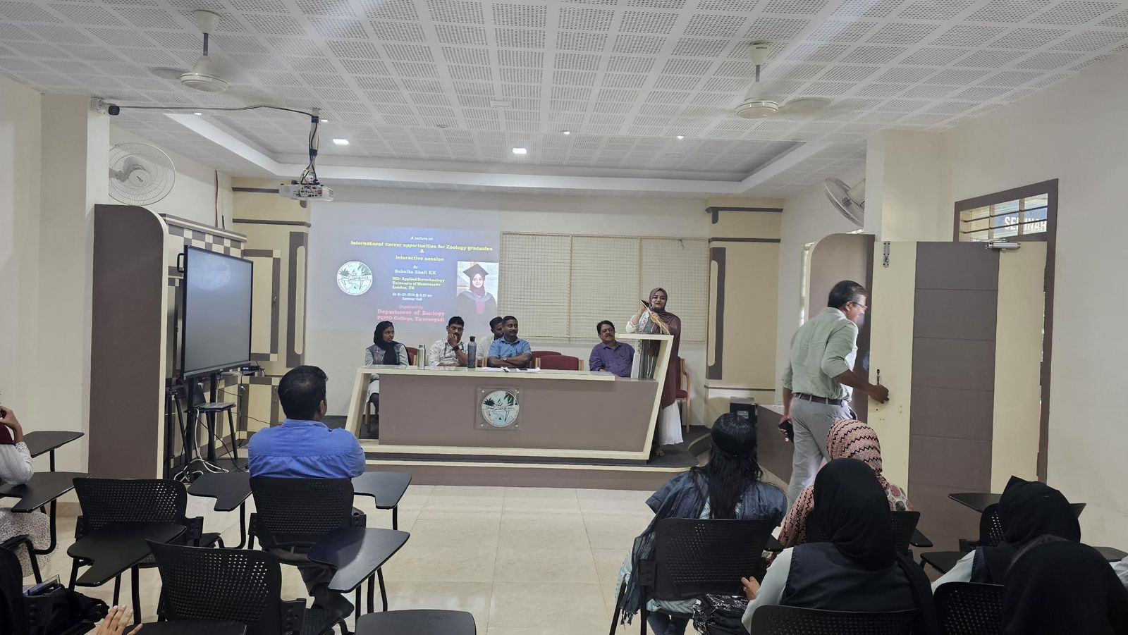 Subaiba speaking at a podium beside a projected lecture poster, with faculty seated at a panel table