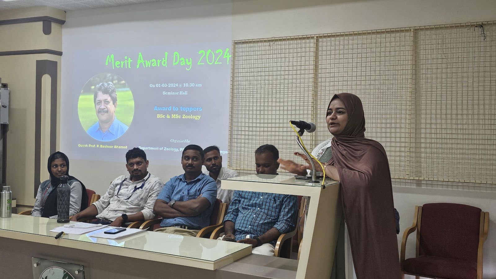 Subaiba speaking at a podium in front of a projected Merit Award Day 2024 slide, with faculty seated alongside