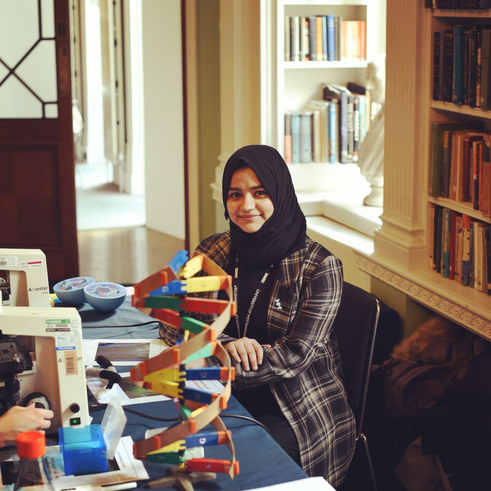 Subaiba seated at an outreach table with a DNA model, microscopes and cell models, in front of bookshelves