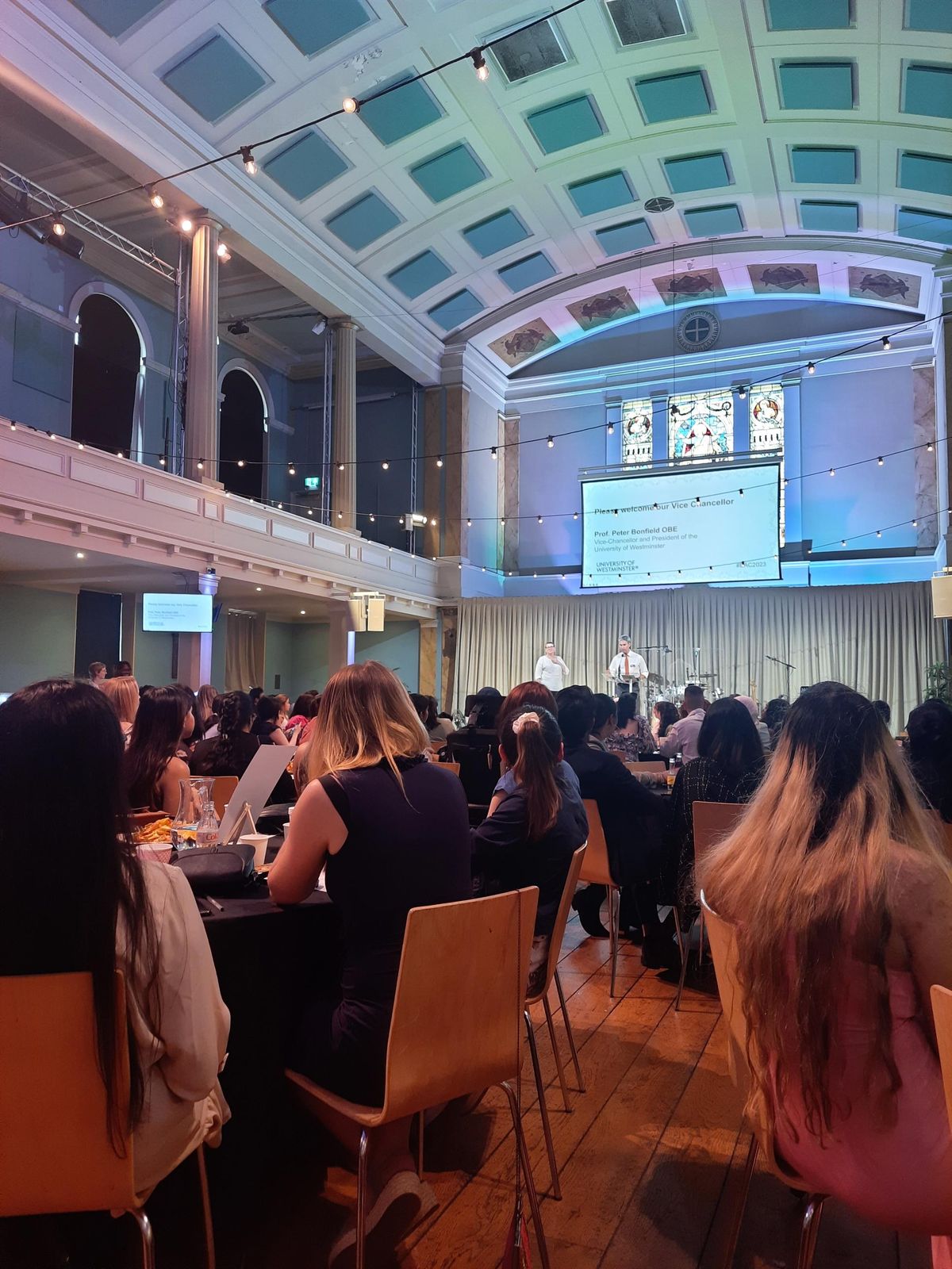 Audience at round tables in a hall with a stained-glass window, facing a screen reading Please welcome our Vice Chancellor